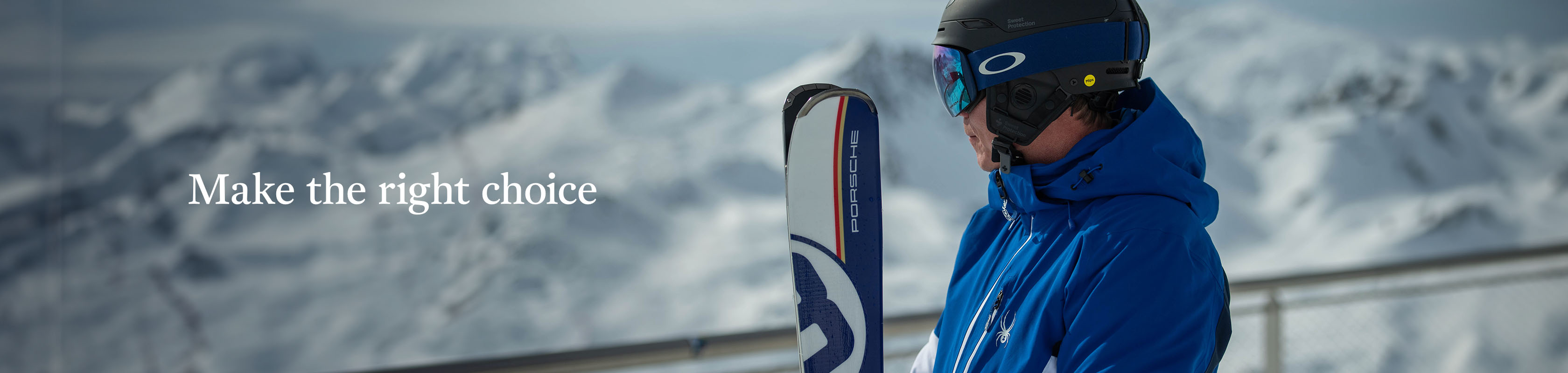 A man in a blue jacket and ski helmet with ski goggles is holding his skis together, somewhere in a ski resort.